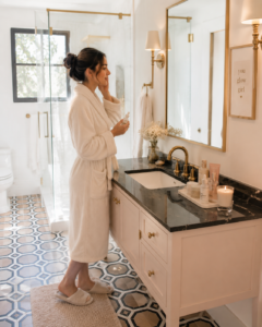 The Must-Have Ultimate Glow-Up Checklist Woman in a white robe applying skincare in a bright luxury bathroom with a minimalist vanity, gold accents, candle, and organized morning routine aesthetic.