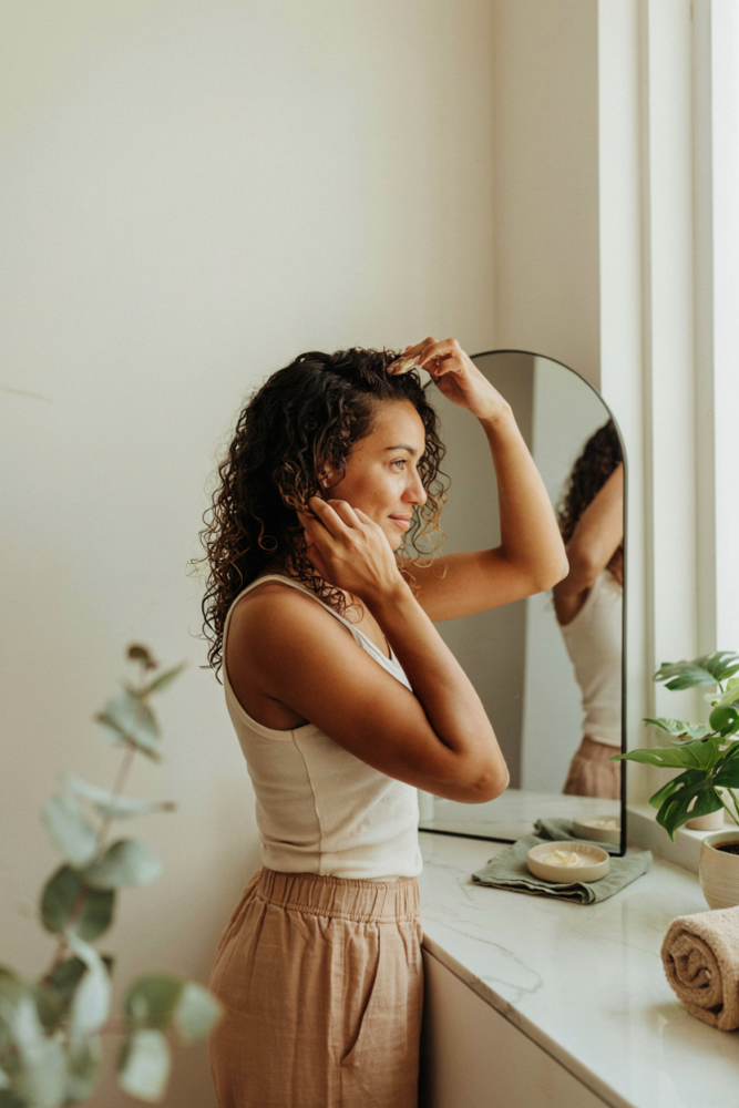 Woman with naturally defined curly hair applying styling cream during her morning curly hair routine in a bright, modern bathroom.